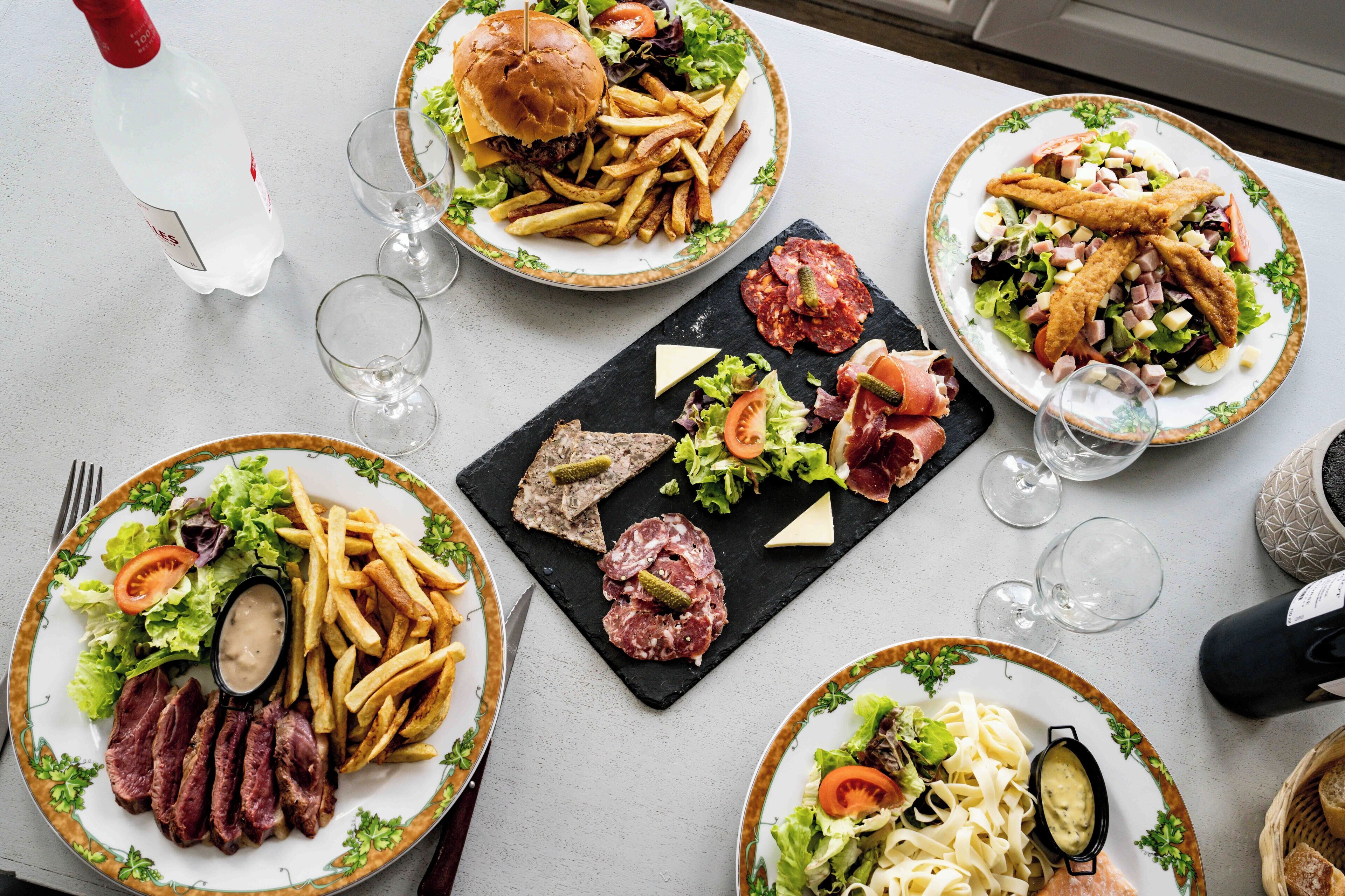 Une table dressée avec divers plats, notamment un hamburger, des frites, une salade et des pâtes, ainsi que des verres à vin et une bouteille d’eau.