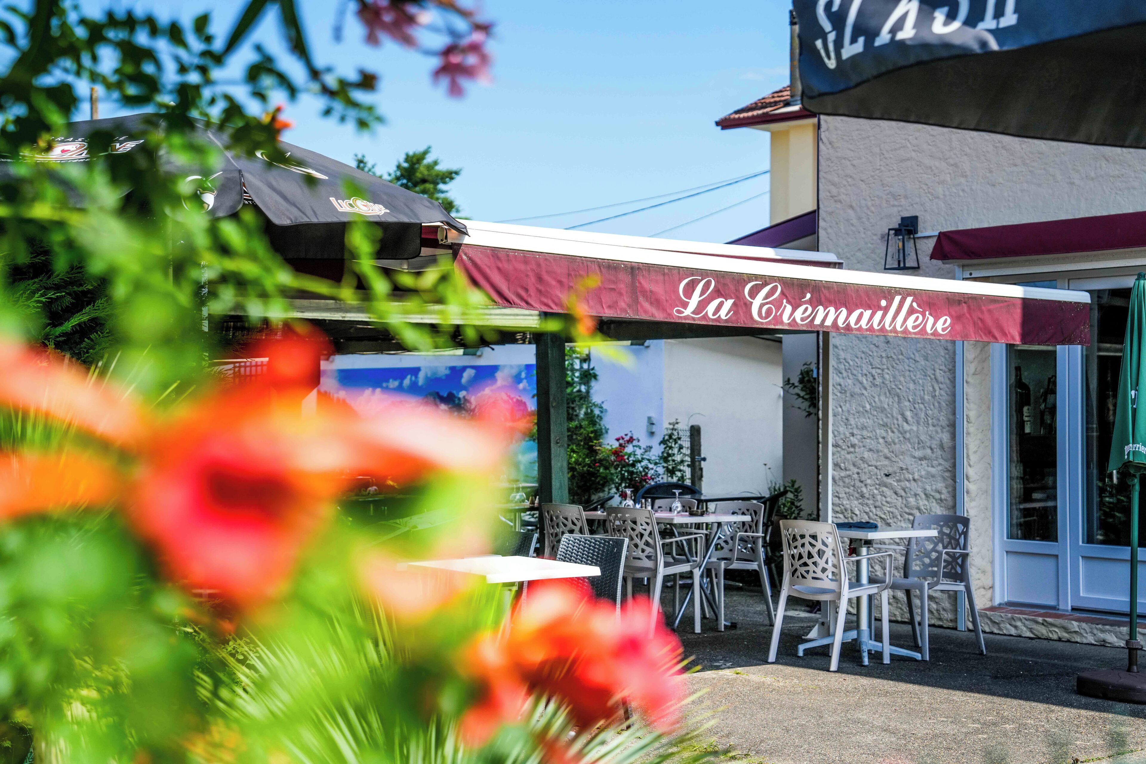 Un café al aire libre con mesas y sillas, un toldo con la inscripción "La Crémailière" y un edificio con un letrero que dice "SLASH".