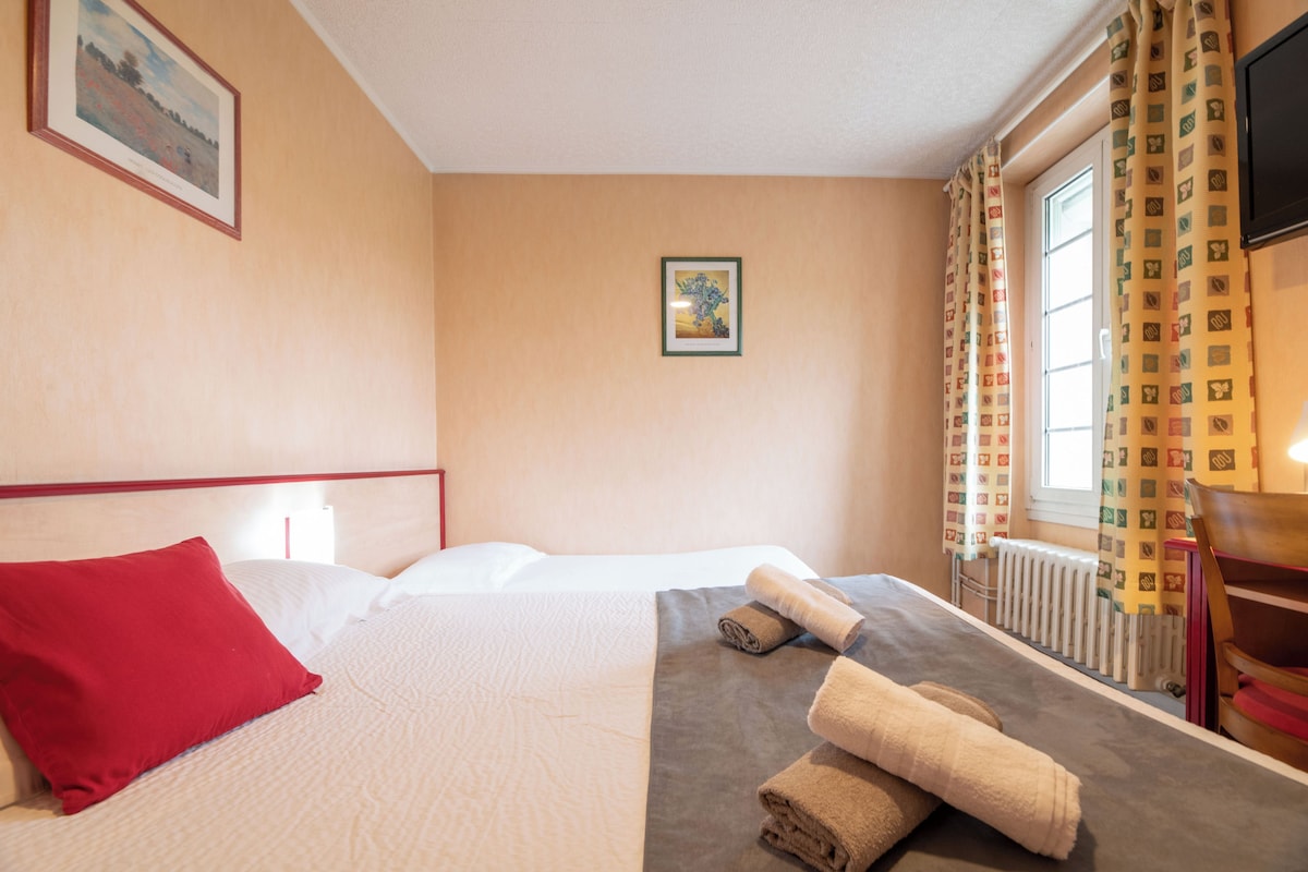 A hotel room with a bed, a red pillow, a framed picture on the wall, a window with curtains, and a radiator.