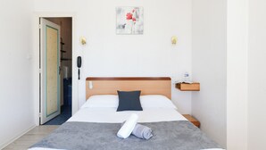 Een net opgemaakt bed met een opgerolde handdoek, een nachtkastje met een fles en een schilderij aan de muur.