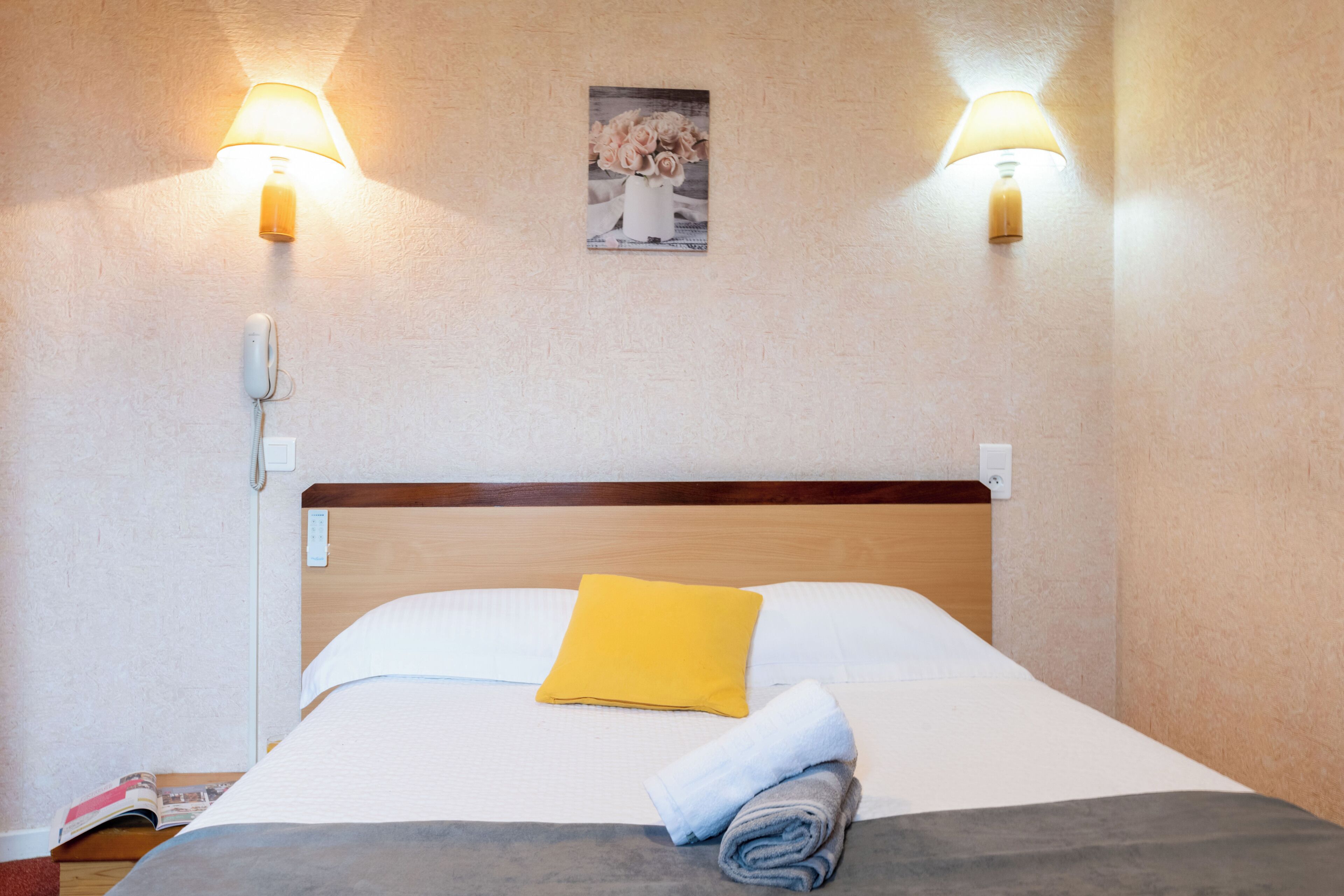Una cama bien hecha con un almohábil amarillo, una toalla doblada y un cuadro en la pared.
