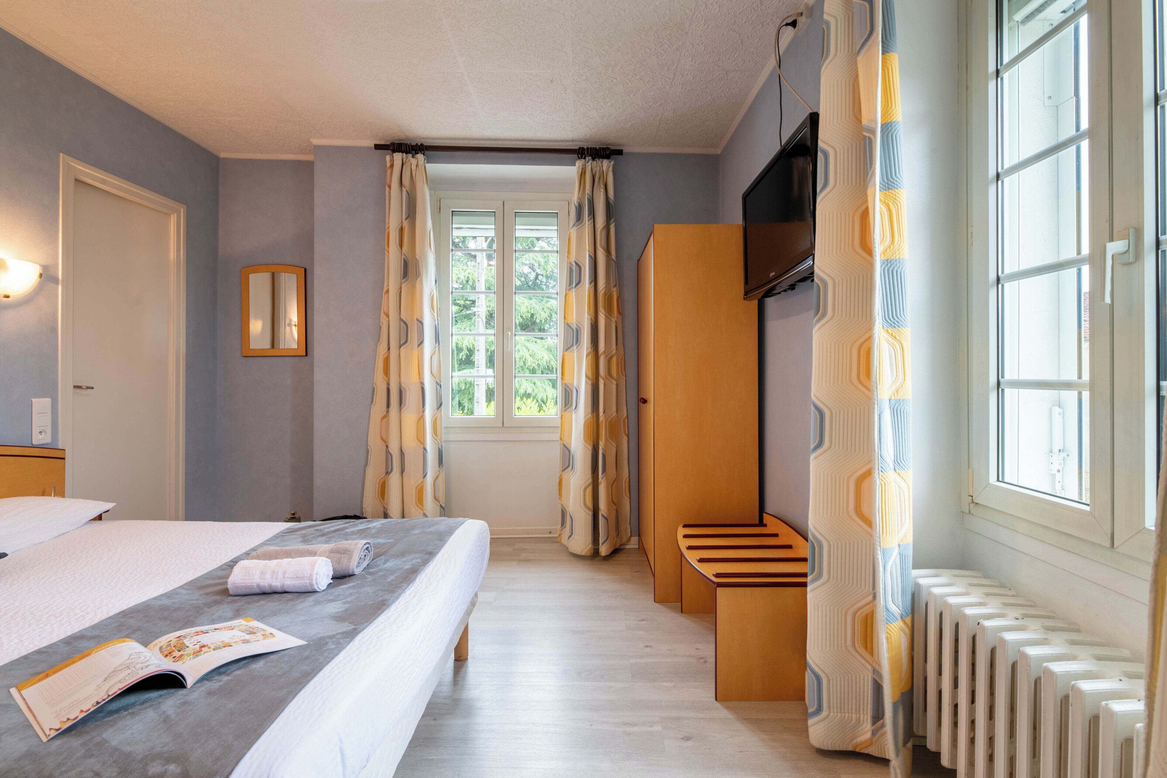 Une chambre d’hôtel avec un lit, une fenêtre avec des rideaux, un radiateur et un petit banc en bois.