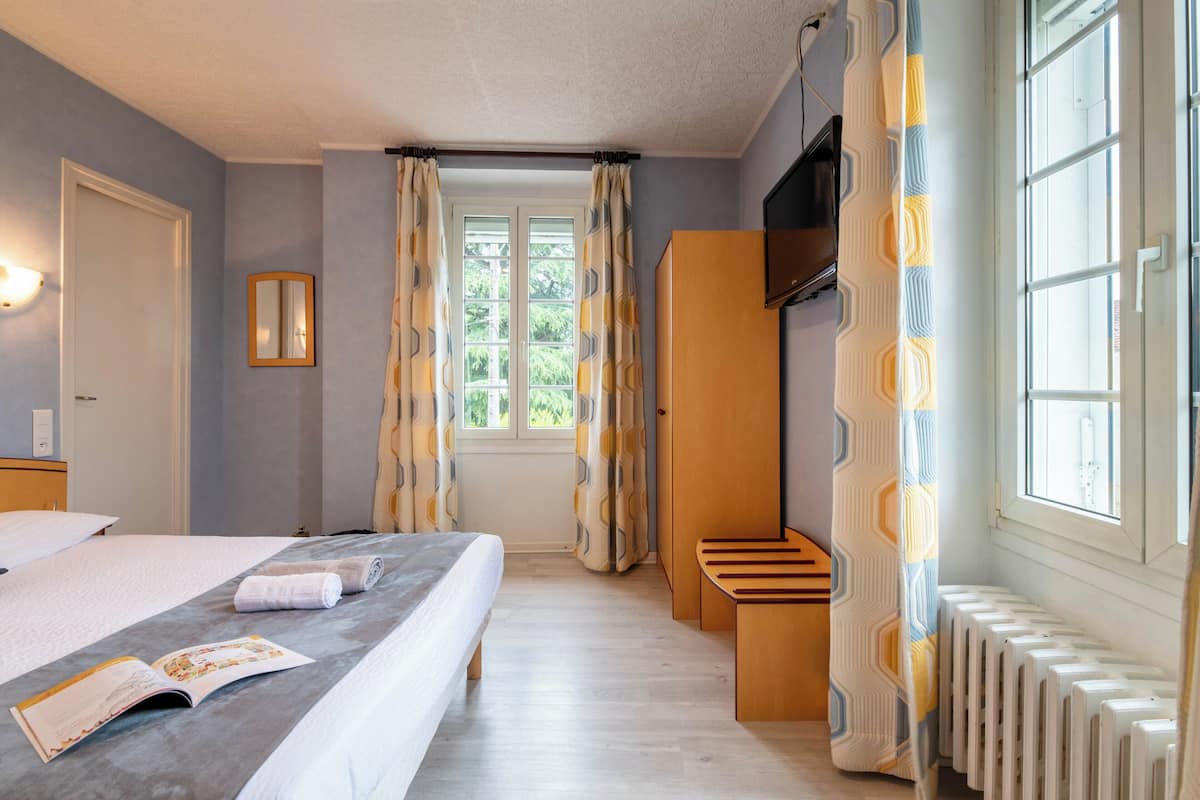 Une chambre d’hôtel avec un lit, une fenêtre avec des rideaux, un radiateur et un petit banc en bois.