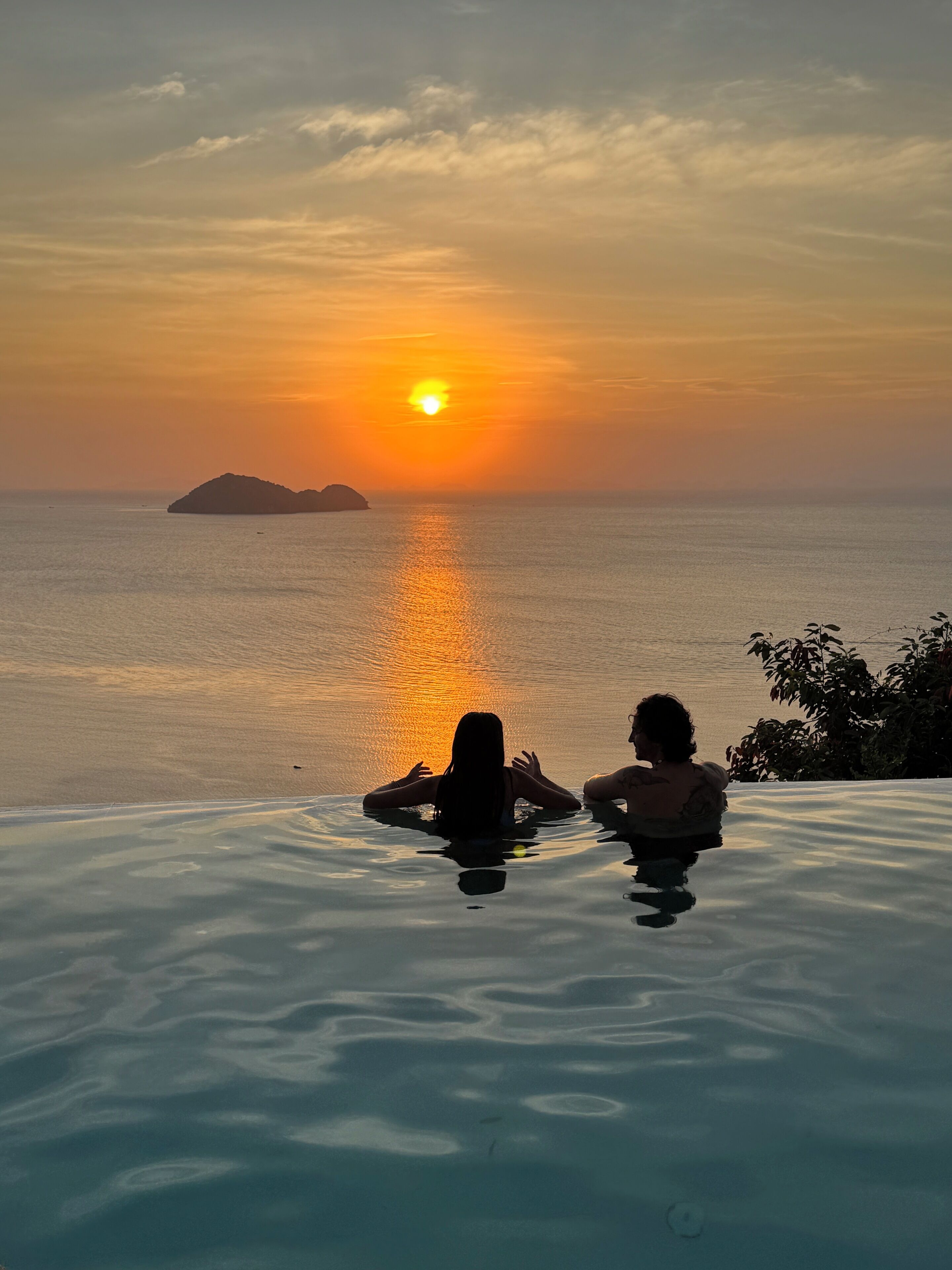 Um pôr do sol sobre um mar calmo, com duas pessoas em uma piscina infinita.