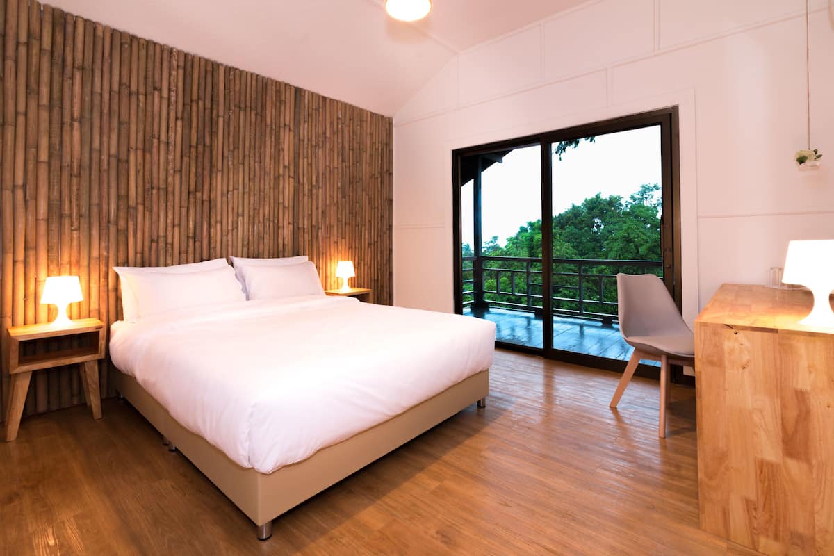 A modern bedroom with a large bed, wooden headboard, two bedside tables with lamps, a wooden desk, and a large sliding glass door leading to a balcony with a view of greenery.