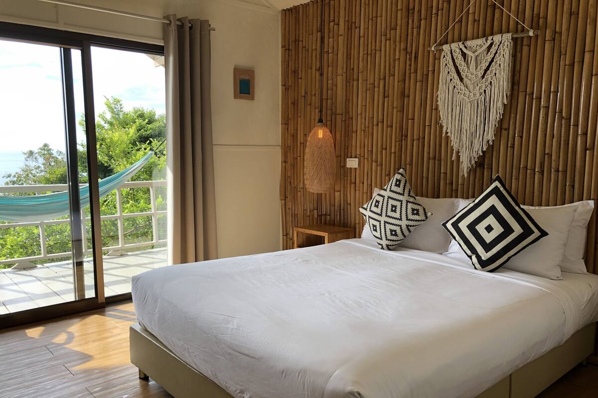 A bedroom with a large bed, a view of greenery through a sliding door, and a bamboo wall.