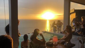 A sunset view from a poolside area with people seated at tables, overlooking the ocean.