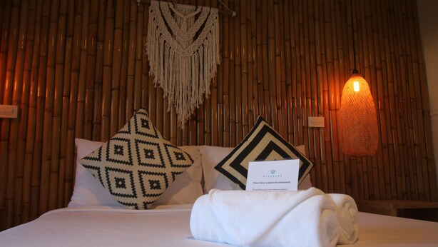 A neatly made bed with a white pillow, a decorative pillow, and a rolled towel. There is a sign on the bed and a wall-mounted light fixture. - Bluerama Resort (Ko Pha-ngan)