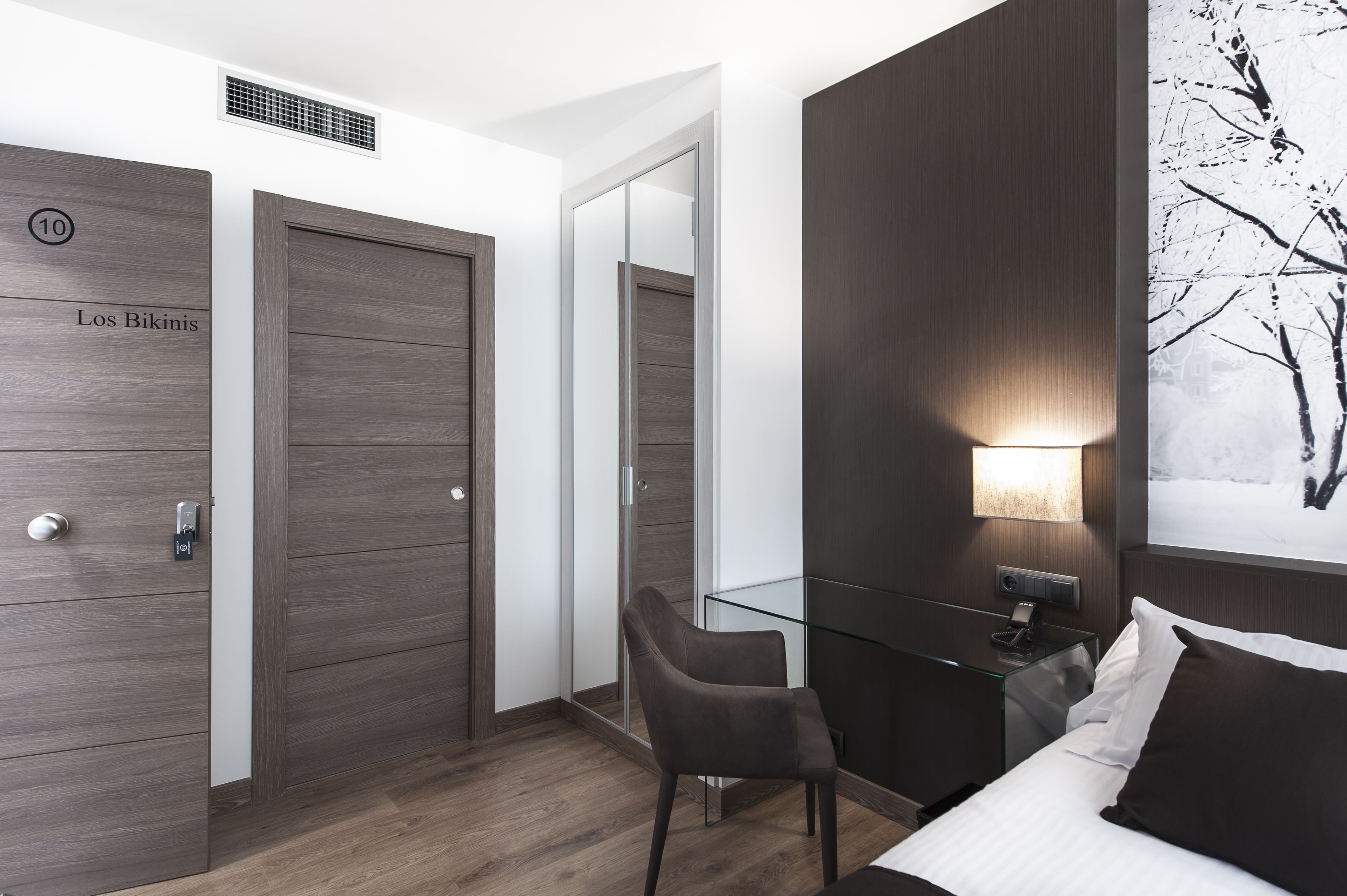 A modern hotel room with a wooden wardrobe, a glass desk, a chair, a bed, and a wall-mounted lamp.