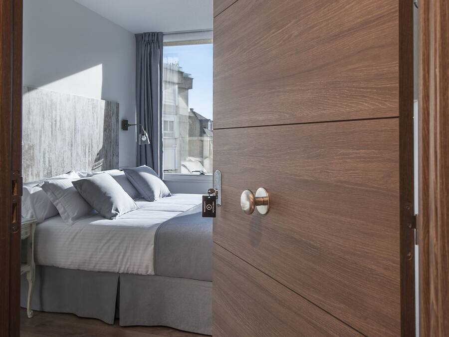 Habitación de hotel con una puerta de madera que dice "La Concha", una cama con ropa blanca, una mesita de noche y vistas a edificios.