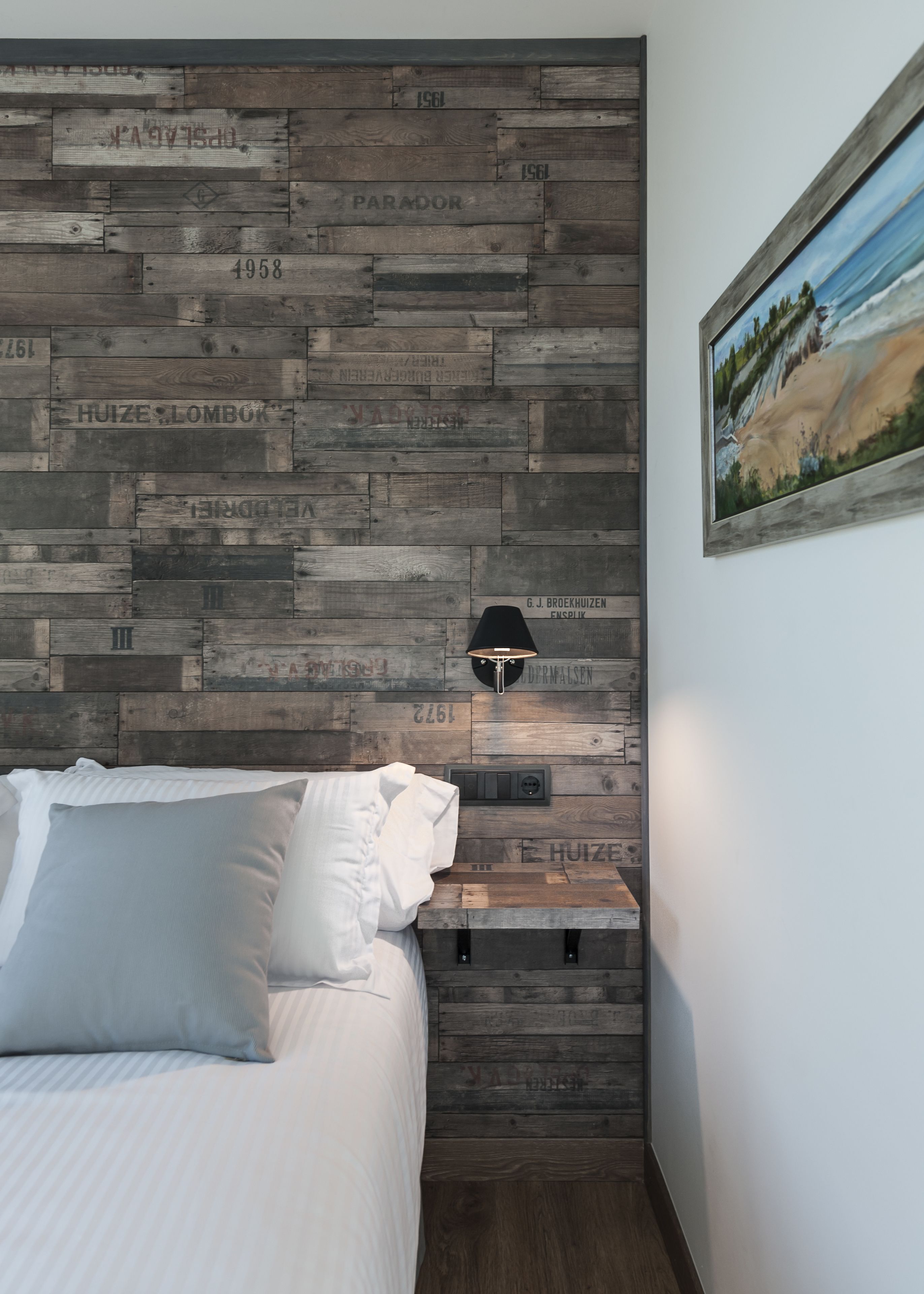 Un dormitorio con cabecera de madera, cama con ropa de cama blanca, mesita de noche con lámpara y un cuadro en la pared.