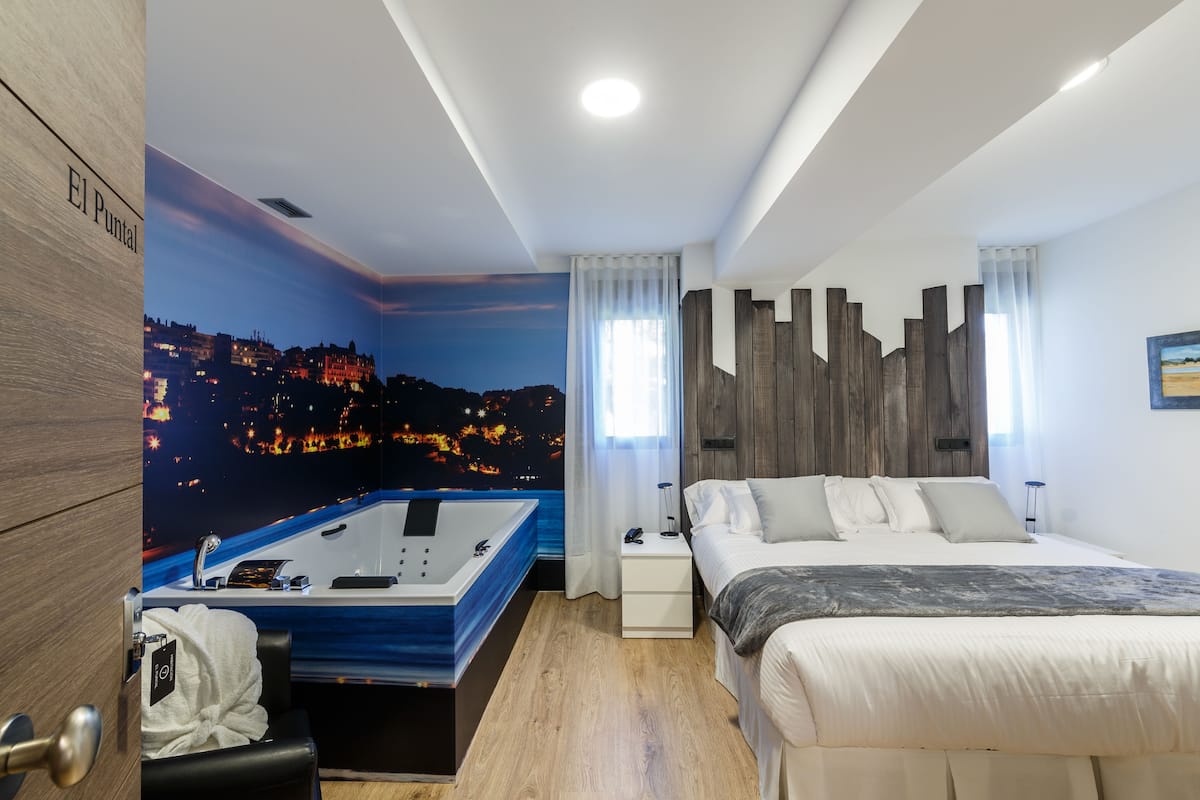A modern hotel room with a cityscape mural, a large bed, a nightstand, a bathroom with a bathtub, and a wooden accent wall.