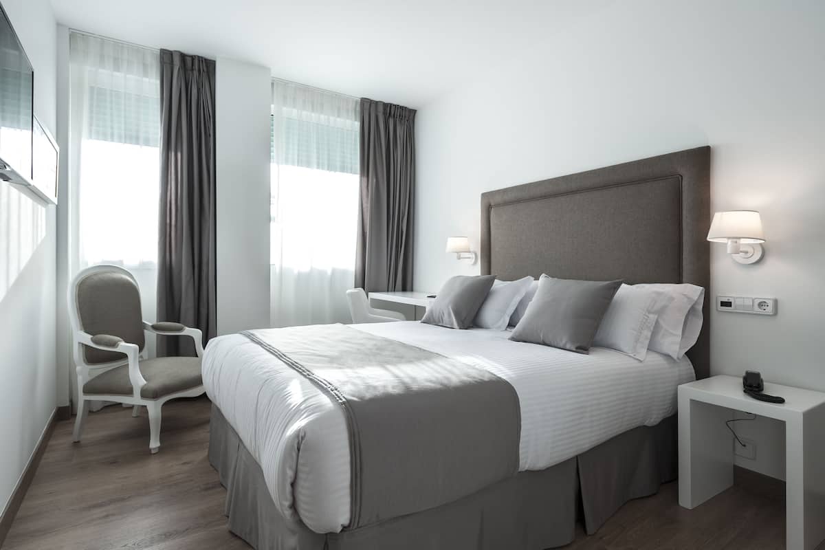 A modern hotel room with a large bed, a chair, a desk, and a nightstand.