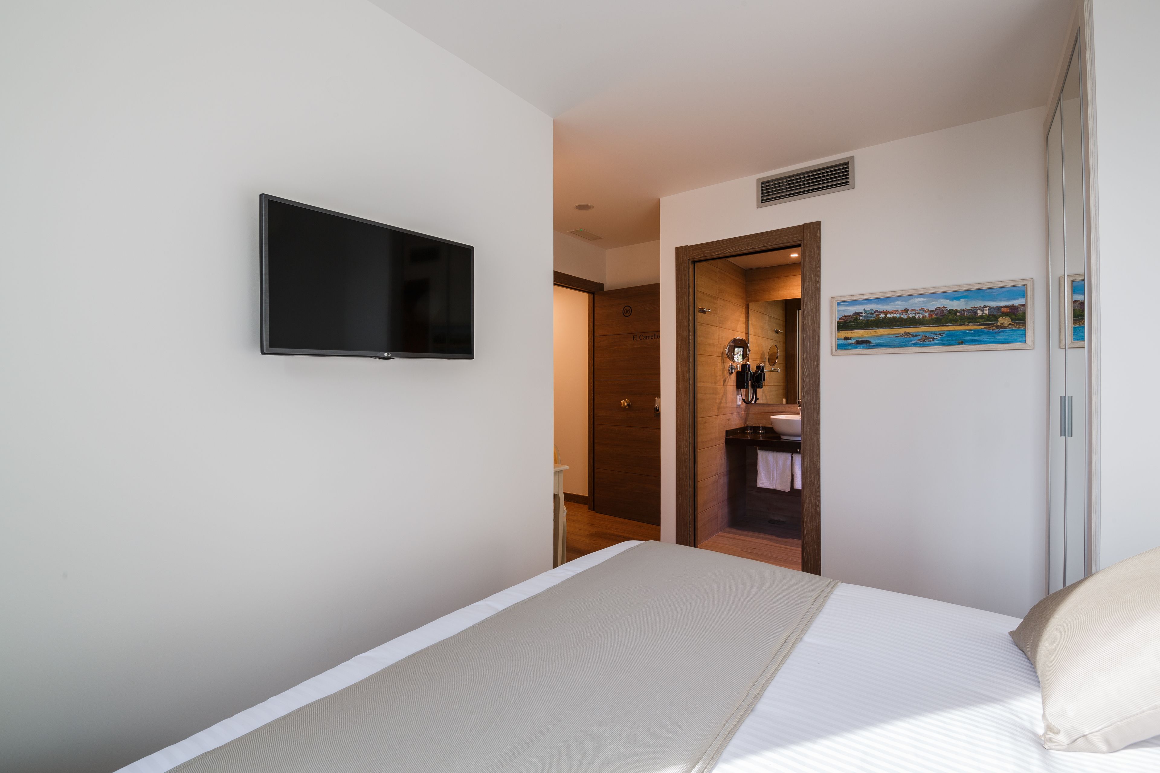 Habitación de hotel con una cama, un televisor montado en la pared, una puerta de madera que da a un baño y un cuadro en la pared.