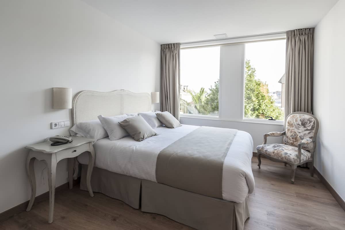 A hotel room with a bed, bedside table, lamp, chair, and a window with curtains.