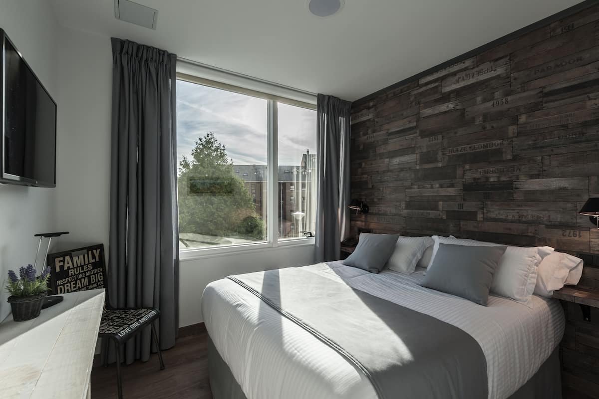 A bedroom with a large bed, a TV mounted on the wall, a window with curtains, and a wooden accent wall.