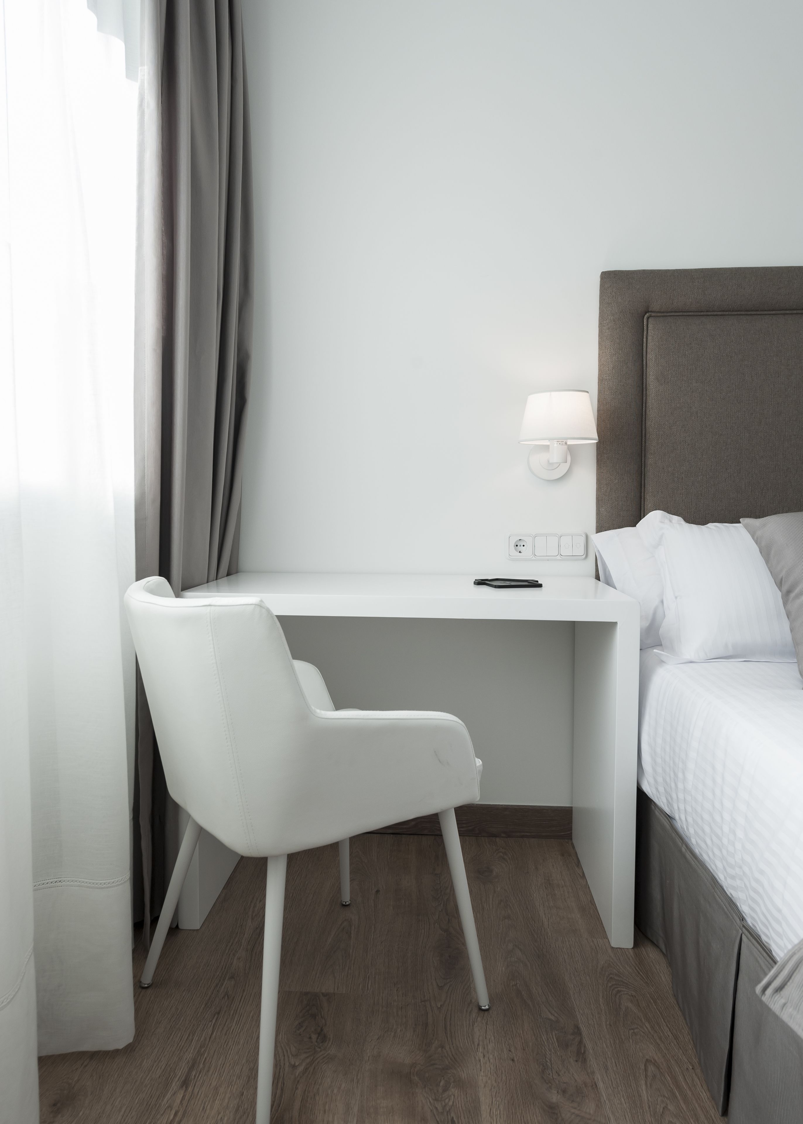 A modern hotel room with a white desk, a white chair, a bed with white bedding, and a grey headboard.