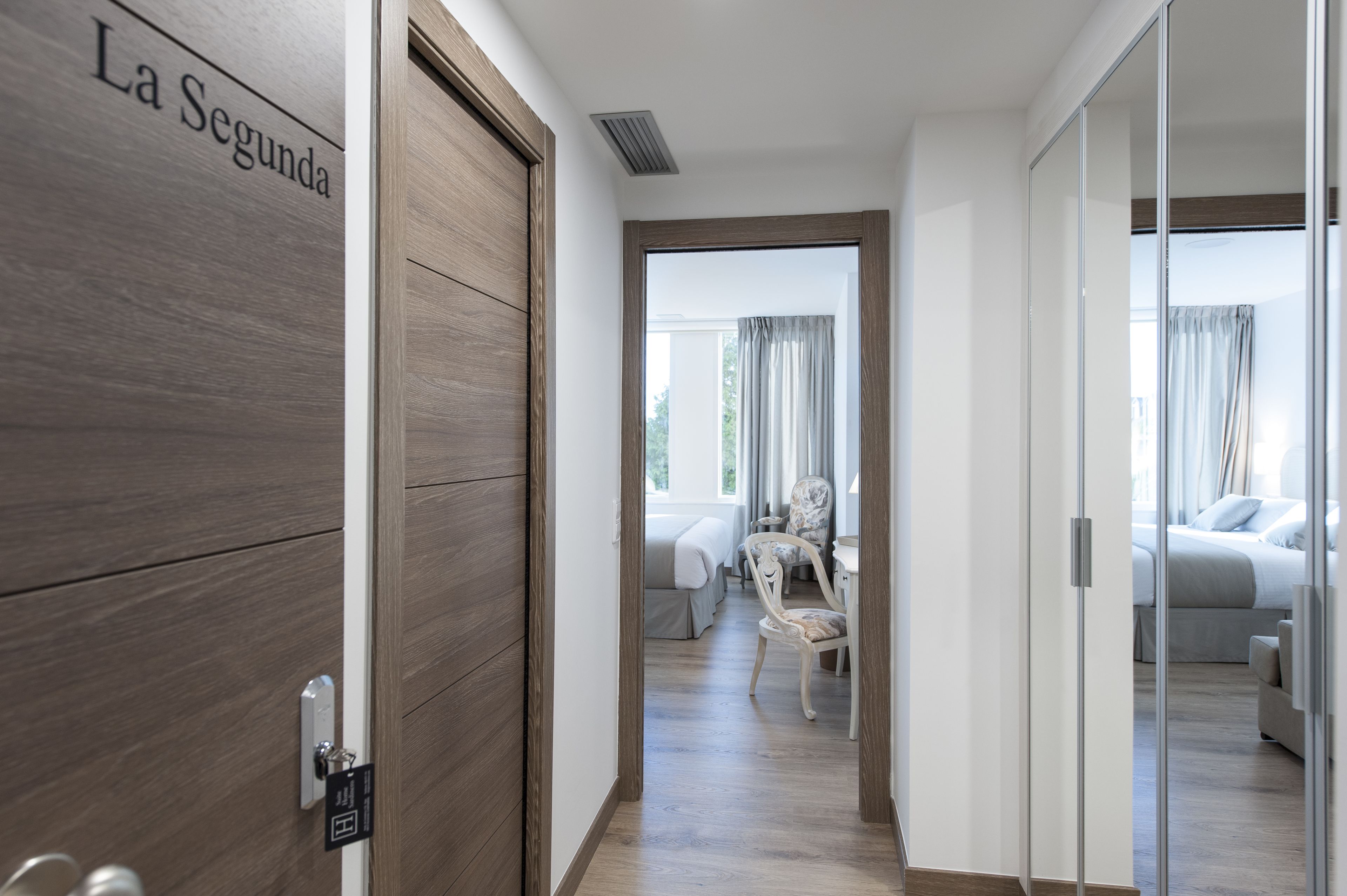 A hotel room with a wooden door labeled "La Segunda," a bed, a chair, and a desk with a lamp.