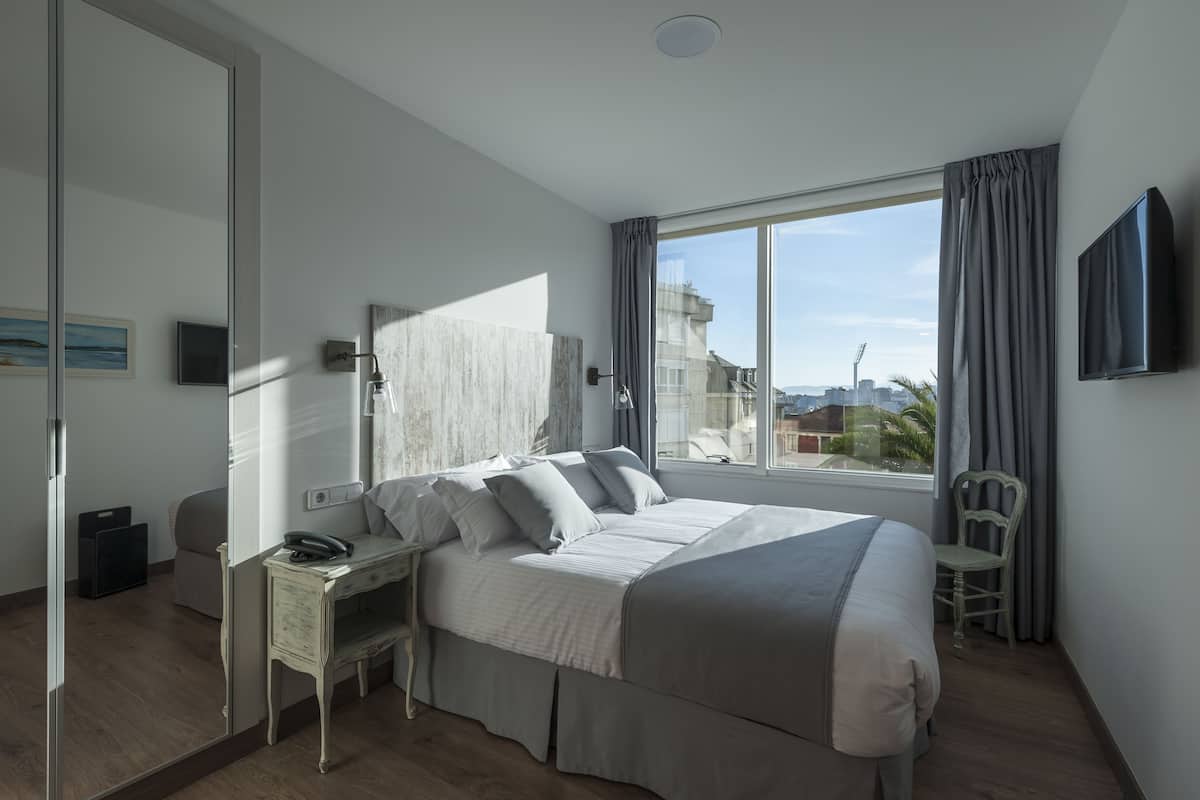 A modern bedroom with a large bed, a desk, and a chair. It features a large window with a view of buildings and a cityscape.