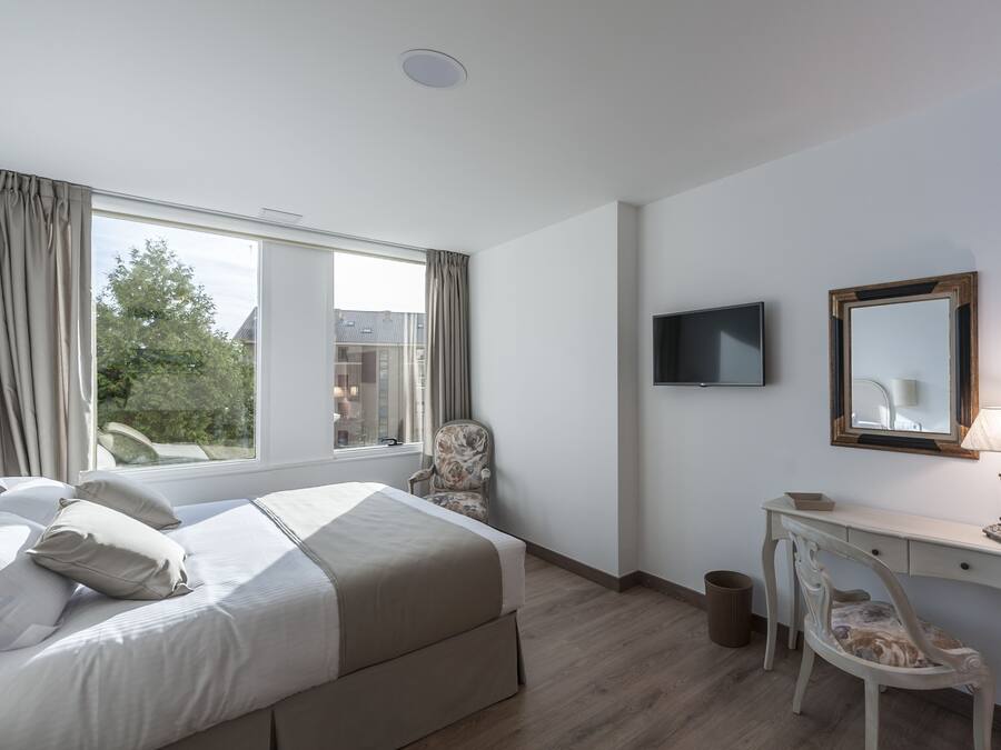 Una habitación de hotel con cama, escritorio, silla, televisor y ventana con cortinas.
