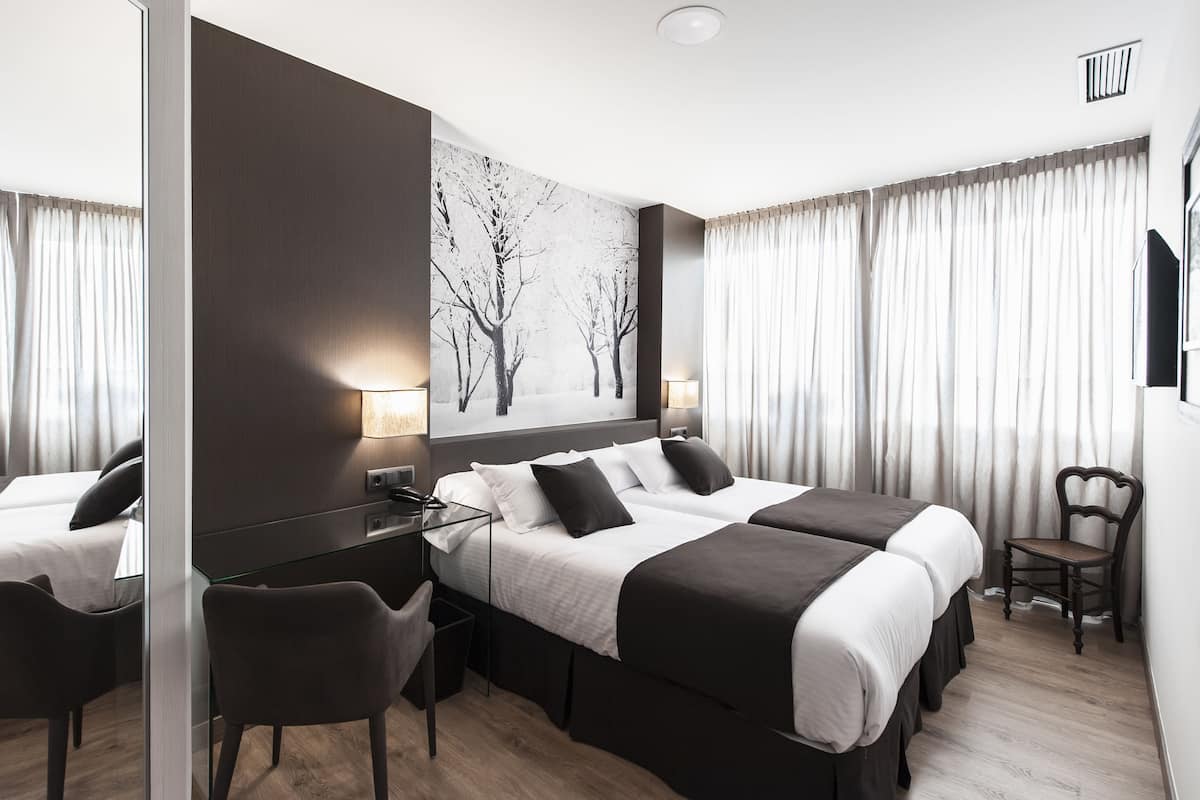 A hotel room with two beds, a desk, a chair, a large window with curtains, and a wall-mounted TV.