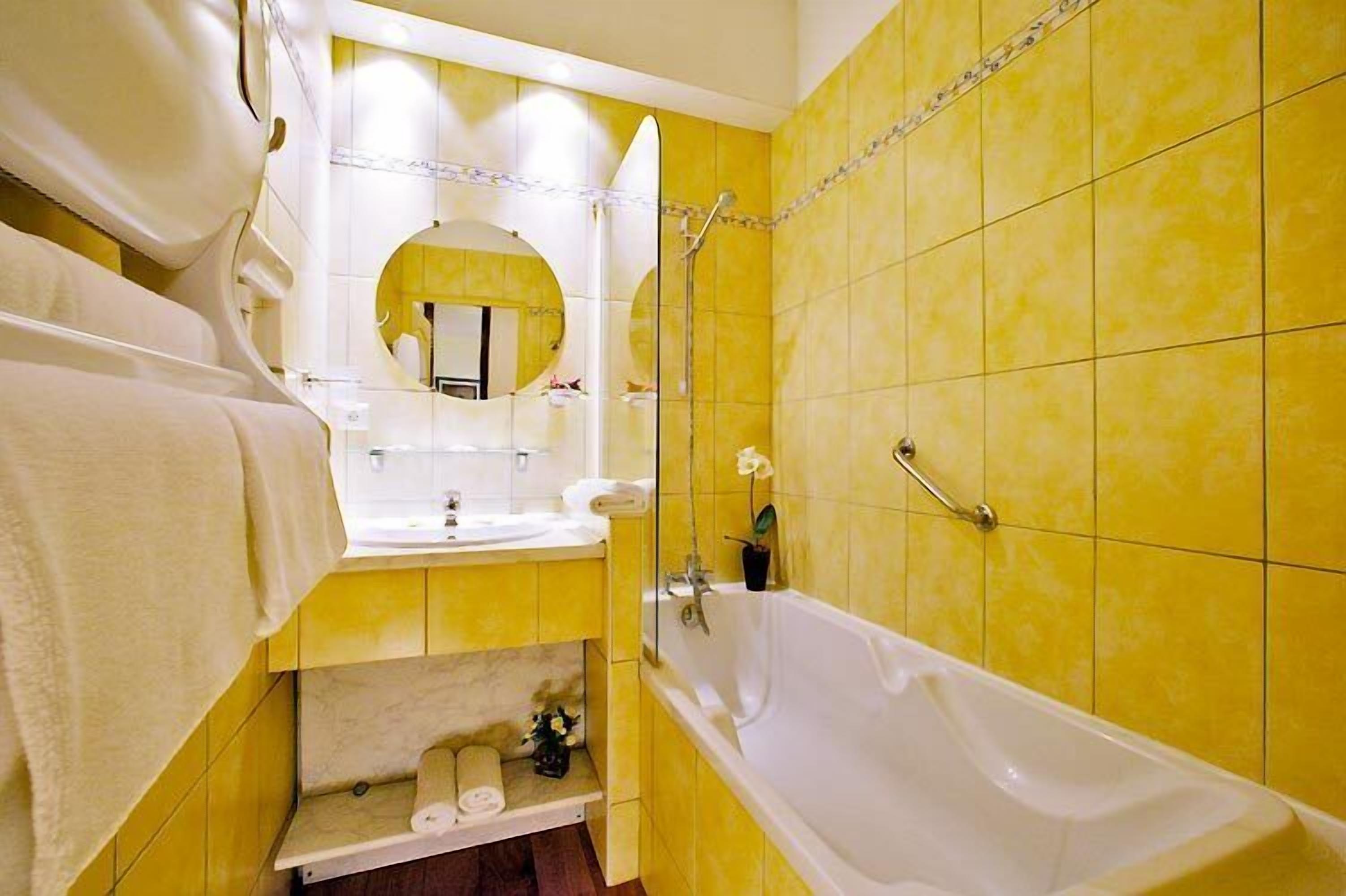 Une salle de bain avec des carreaux jaunes, une baignoire blanche, un lavabo et un miroir doté d’un cadre circulaire.