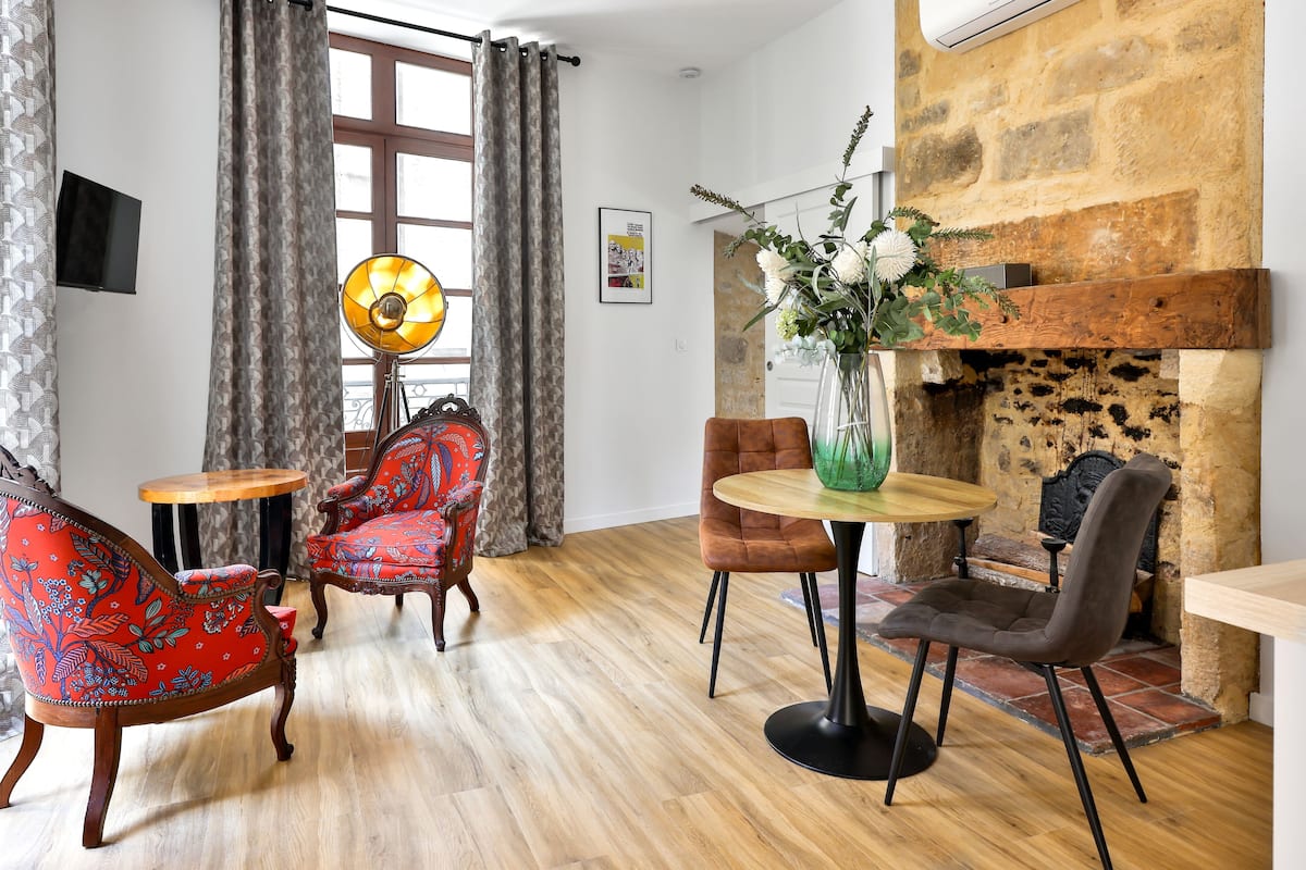 Une pièce dotée d’une cheminée, d’un plancher en bois et d’un mélange de meubles modernes et classiques, notamment un fauteuil rouge à motifs, un fauteuil marron et une table ronde agrémentée d’un vase de fleurs.