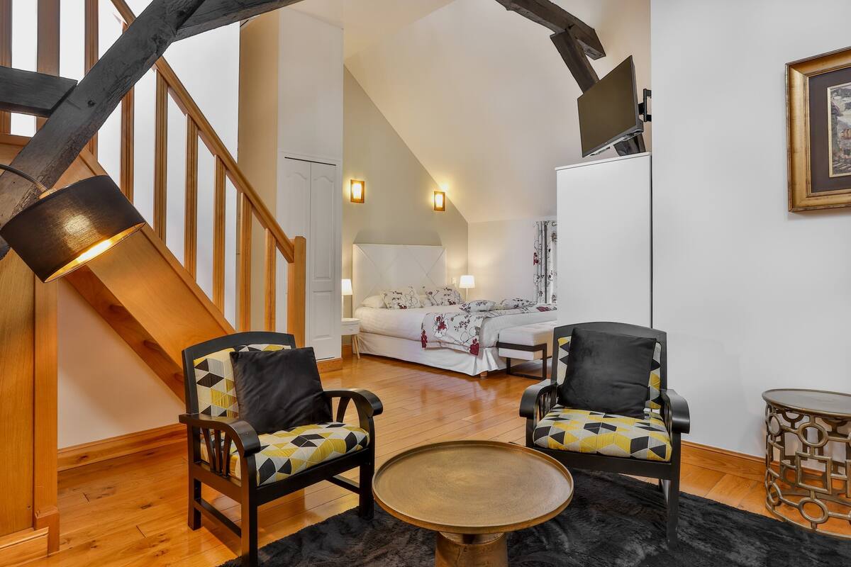 Un salon moderne avec un escalier en bois, deux fauteuils ornés de coussins à motifs, une table basse ronde et un lit recouvert d’une couvre-lit à fleurs.
