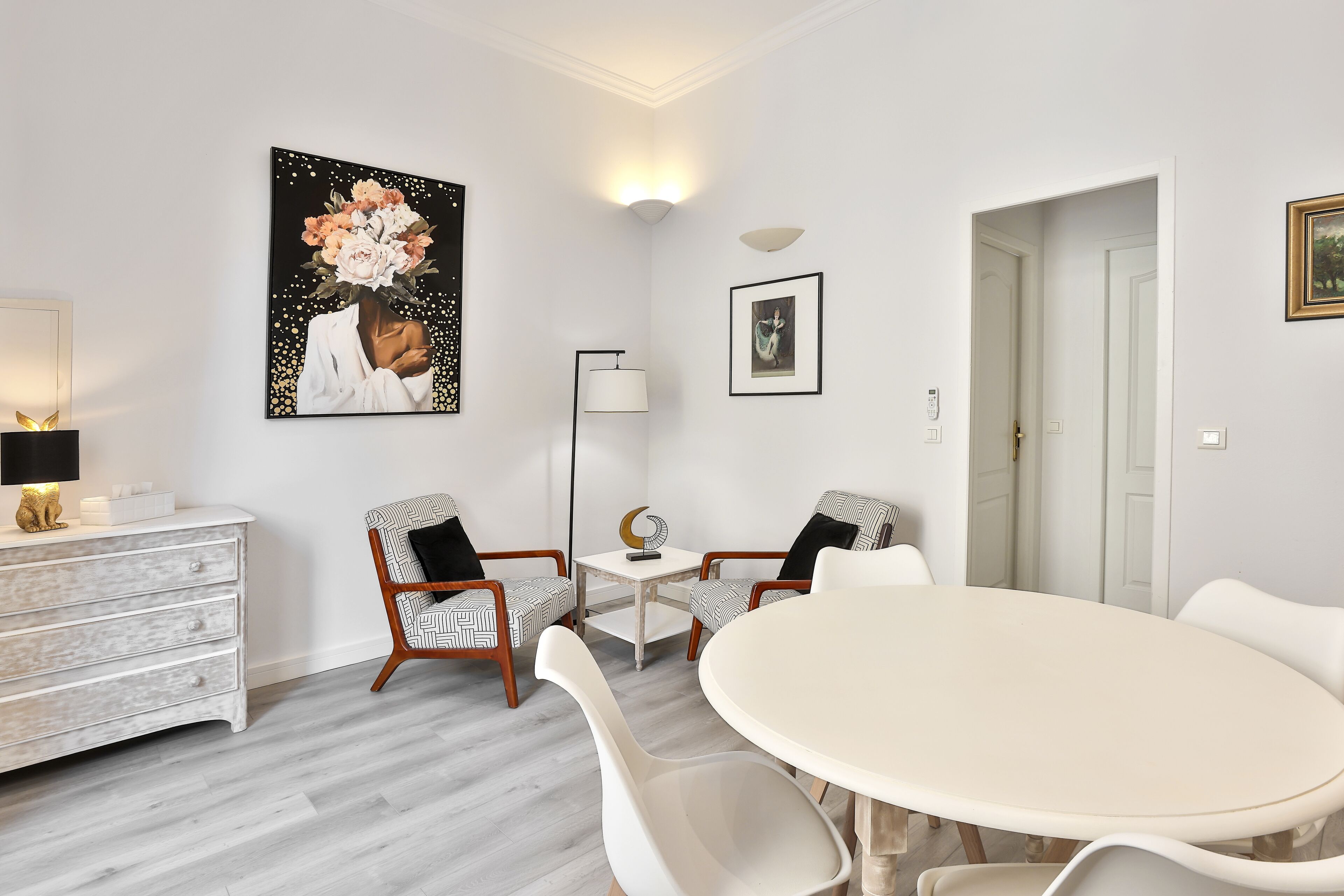 A modern living room with a white round table, two patterned chairs, a white dresser, and a large floral painting on the wall.