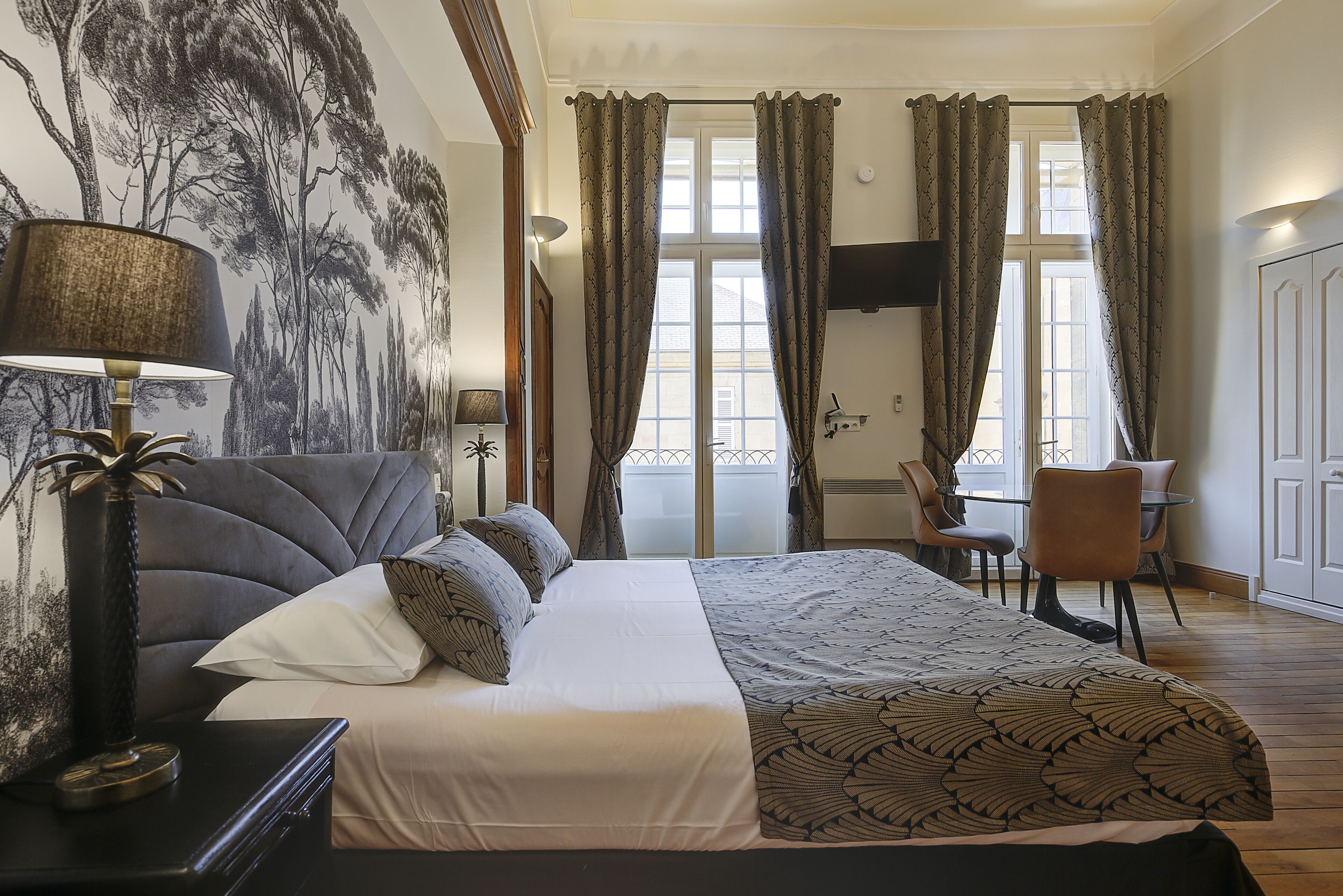 Un dormitorio con cama, escritorio y silla, un ventanal con cortinas y una pared con un mural de un árbol.