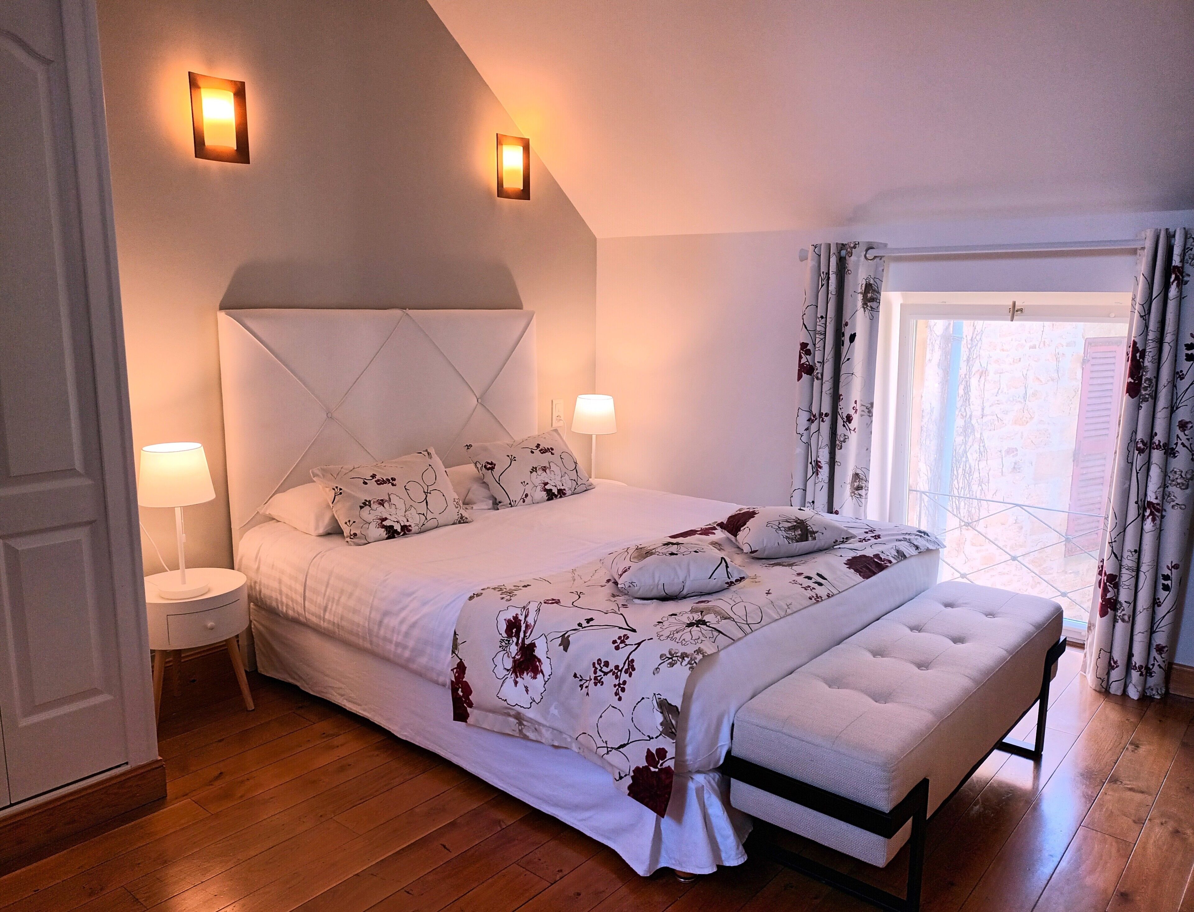 A bedroom with a large bed, two bedside lamps, a window with curtains, and a wooden floor.