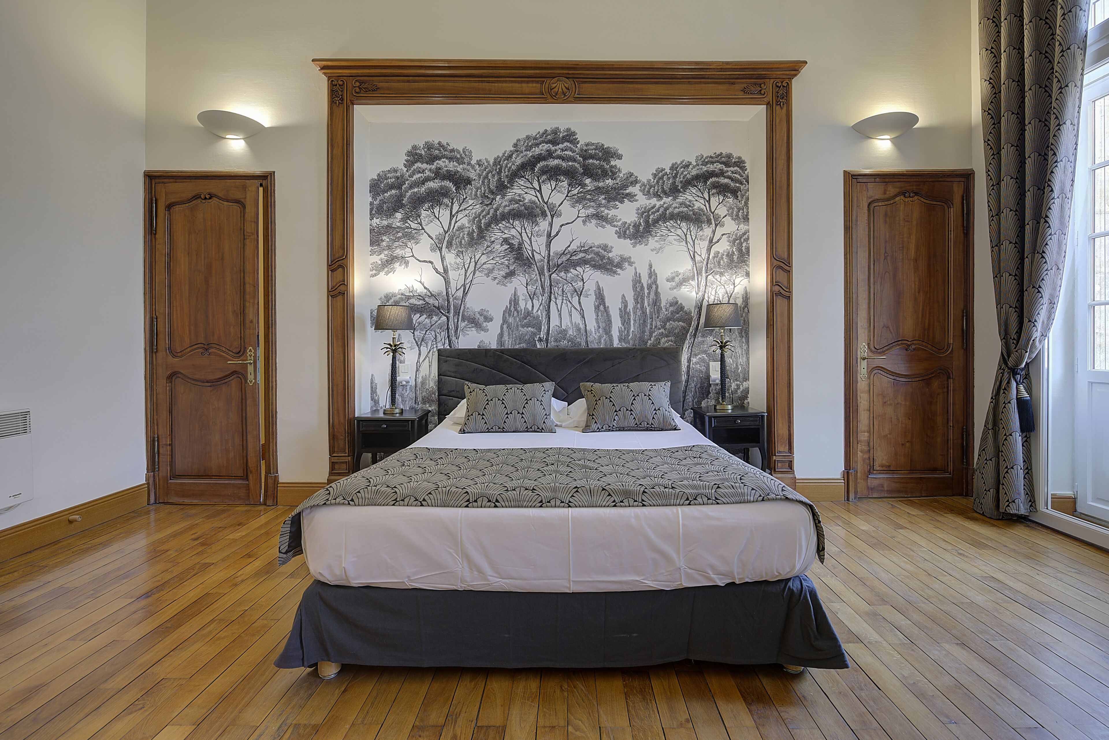 Une chambre à coucher avec un grand lit, deux tables de chevet, une lampe fixée au mur et une porte dotée d’un miroir.