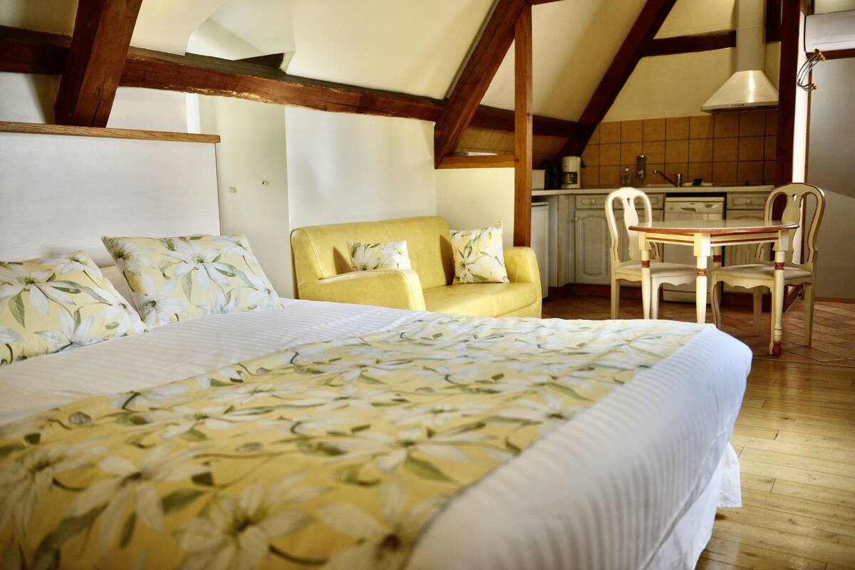 Une chambre avec un grand lit, un fauteuil jaune, une table à manger en bois et, au fond, une cuisine.
