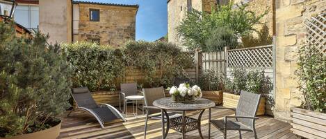 Une terrasse en bois avec une table ronde et des chaises, entourée de plantes en pot et d’un mur de pierre.