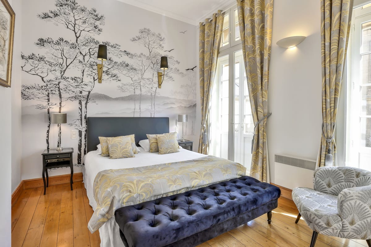 Une chambre à coucher avec un grand lit, un banc, une chaise, une table de chevet et une fresque murale.