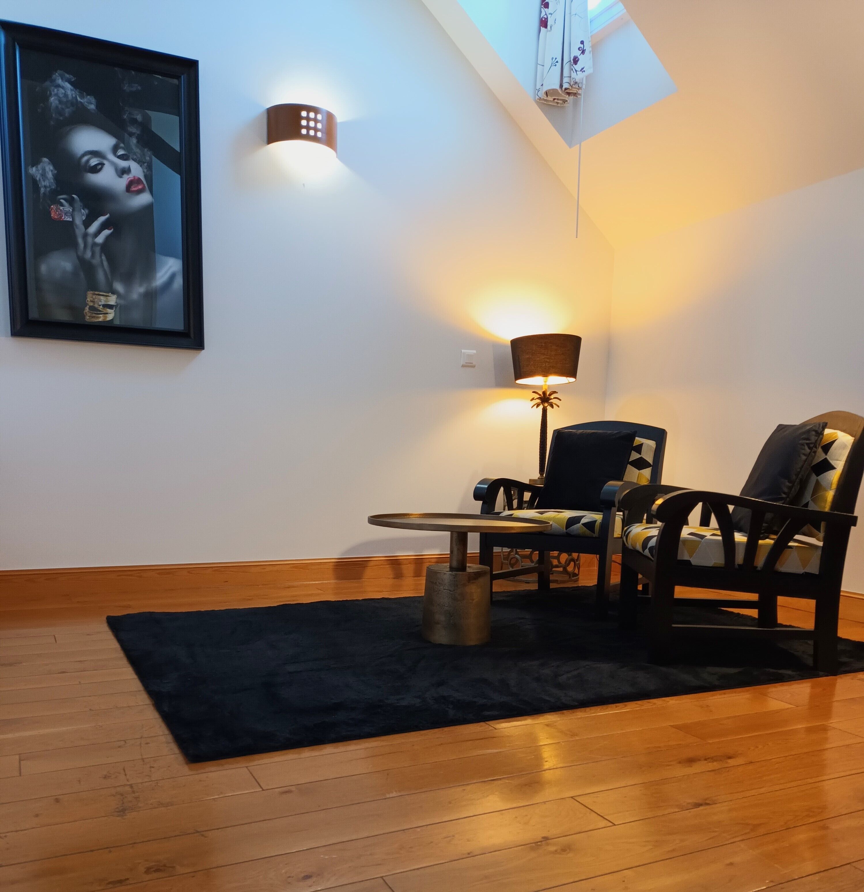 A room with a wooden floor, a black rug, a table, two chairs, a lamp, a framed picture on the wall, and a skylight.