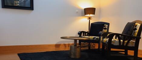 A room with a wooden floor, a black rug, a table, two chairs, a lamp, a framed picture on the wall, and a skylight.