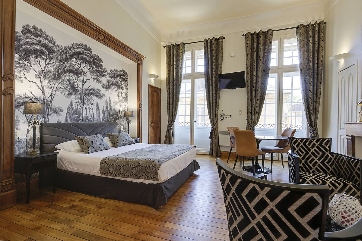 Une chambre avec un grand lit, un bureau et une chaise. Un mur est décoré d’une fresque et il y a une fenêtre avec des rideaux.