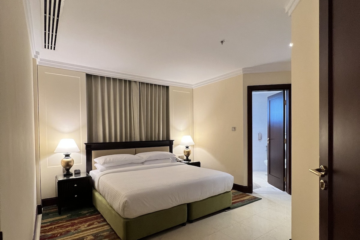 A hotel room with a large bed, two bedside tables with lamps, a headboard, a door, and a small bench.