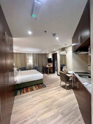 Quarto de hotel com cama, escrivaninha, cadeira, pequeno espaço de cozinha e banheiro.