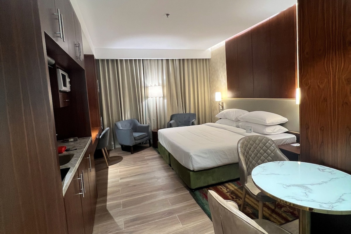 A hotel room with a bed, a desk, a chair, a small table, and a wardrobe.