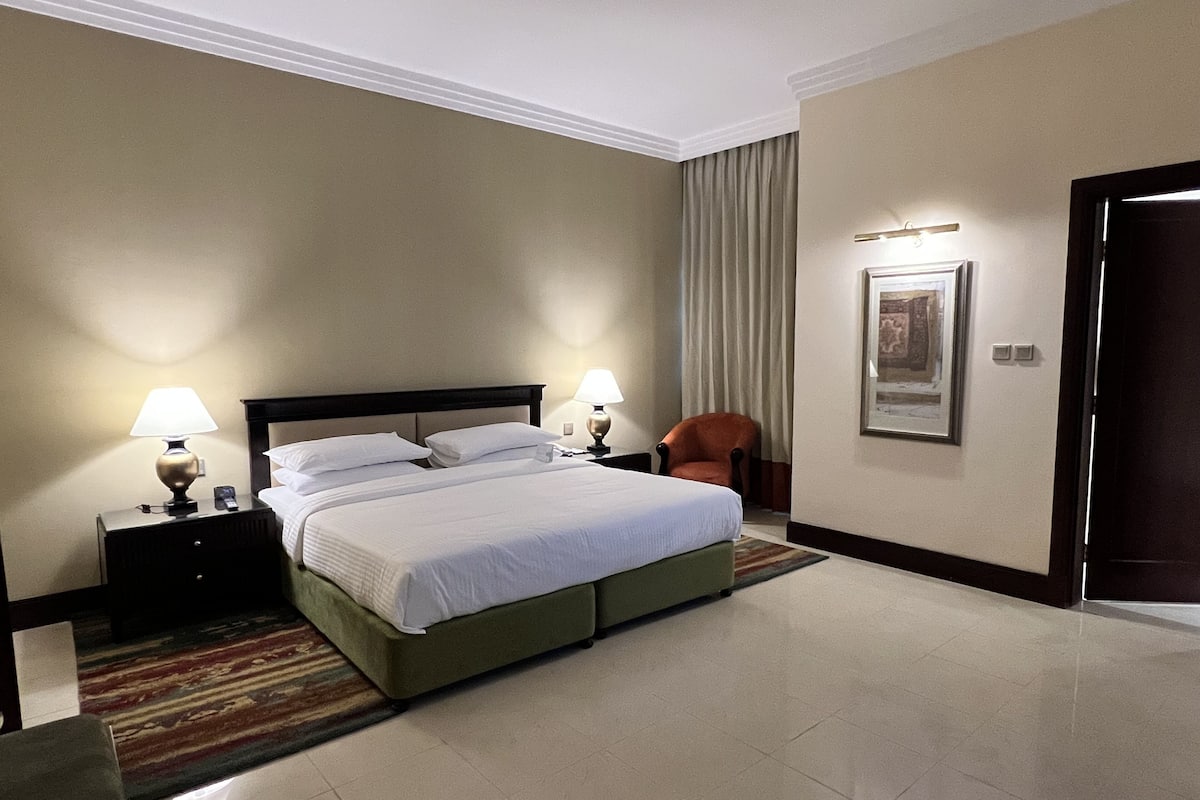 A hotel room with a large bed, two bedside tables with lamps, a chair, a framed picture on the wall, and a door.