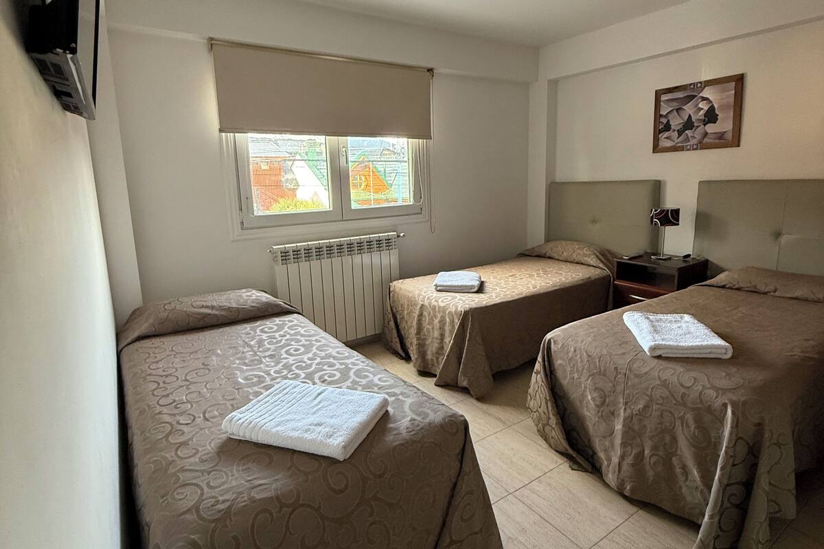 A hotel room with two single beds, a window with blinds, a radiator, a flat-screen TV mounted on the wall, and a framed artwork on the wall.