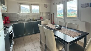 Een keuken met een eethoek, houten balken in het plafond, twee ramen met uitzicht op de bergen, en een rode waterkoker op het aanrecht.
