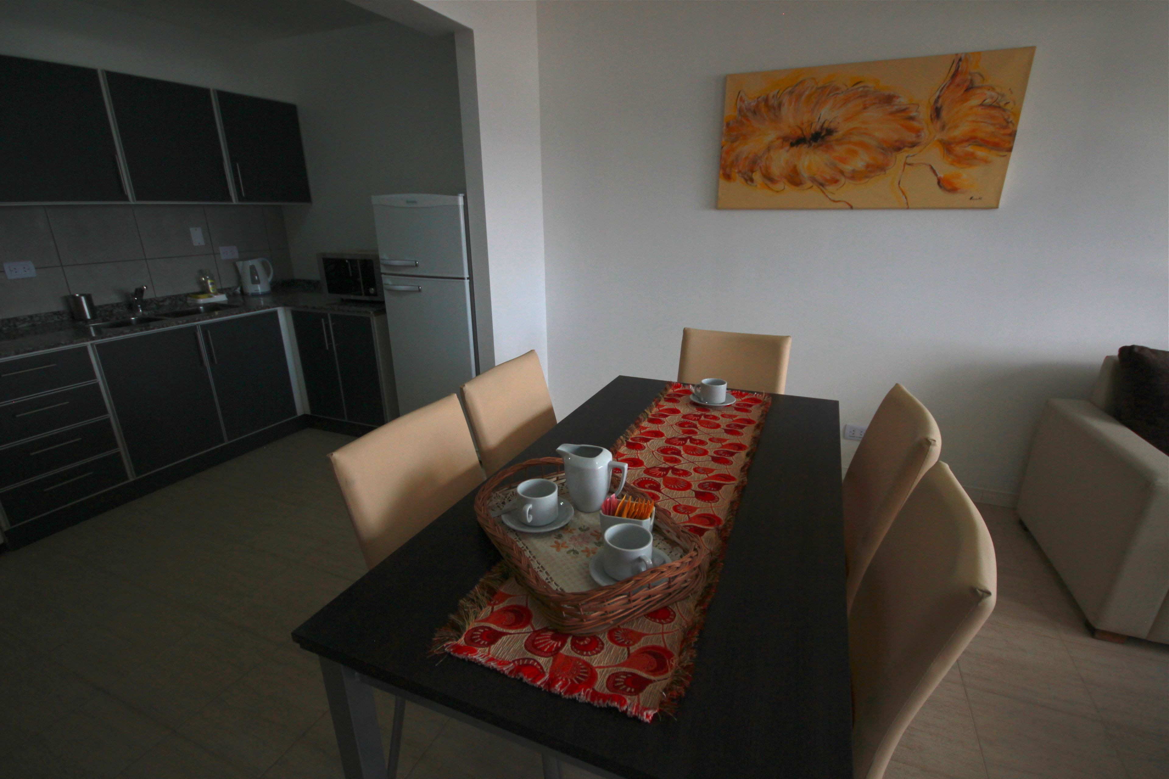 A dining area with a table set for two, a kitchen in the background, and a painting on the wall.
