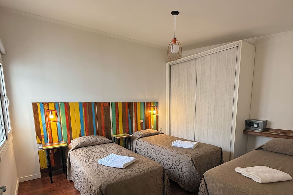 A hotel room with three beds, a wooden floor, a colorful striped wall, and a modern pendant light.