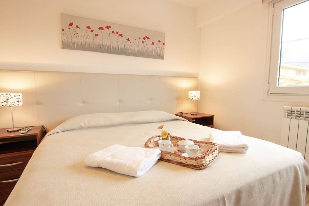 A neatly made bed with a tray of tea and a basket, two bedside tables with lamps, and a painting on the wall.