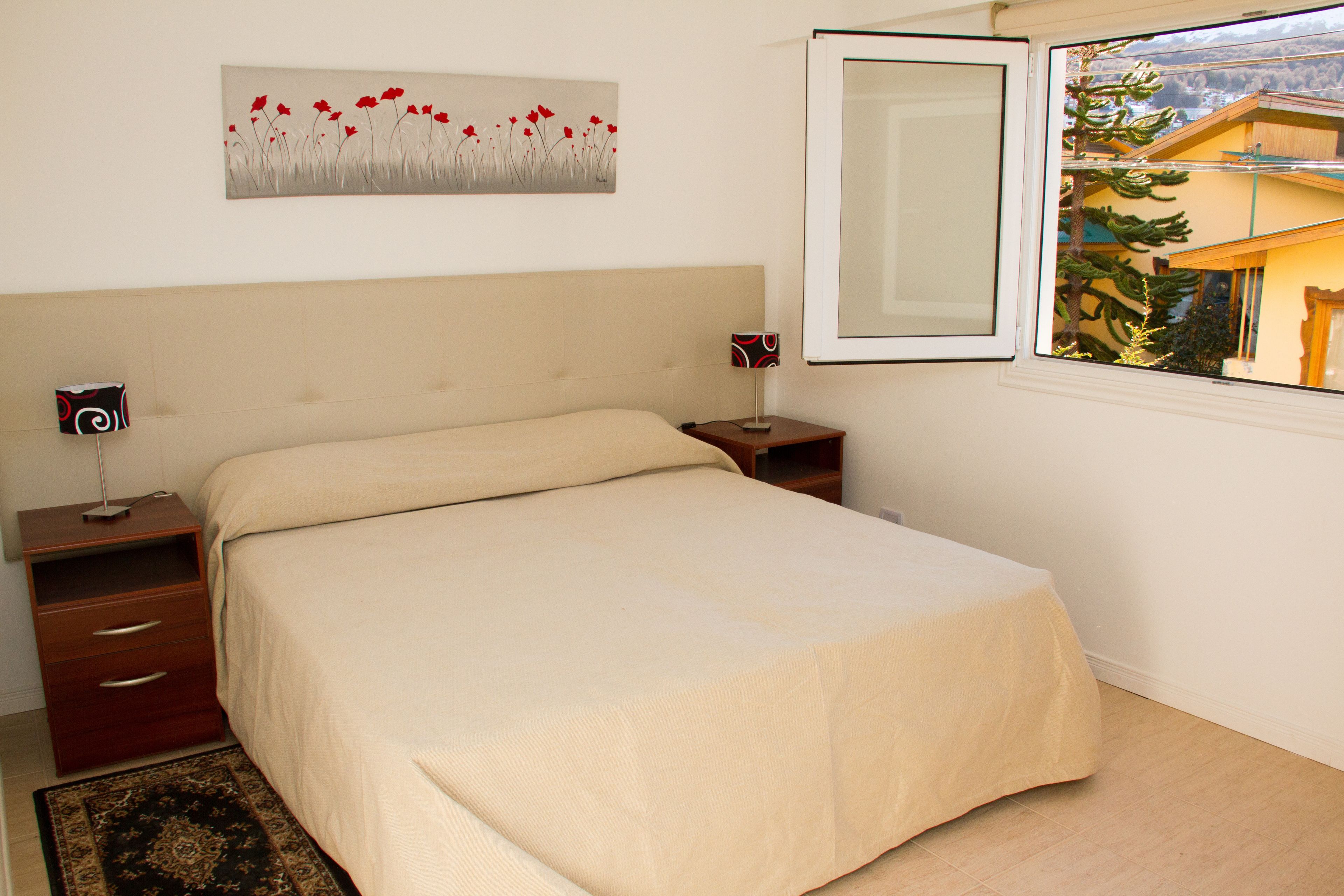 A bedroom with a bed, a nightstand, a lamp, a painting, and a window with a view of trees and a building.