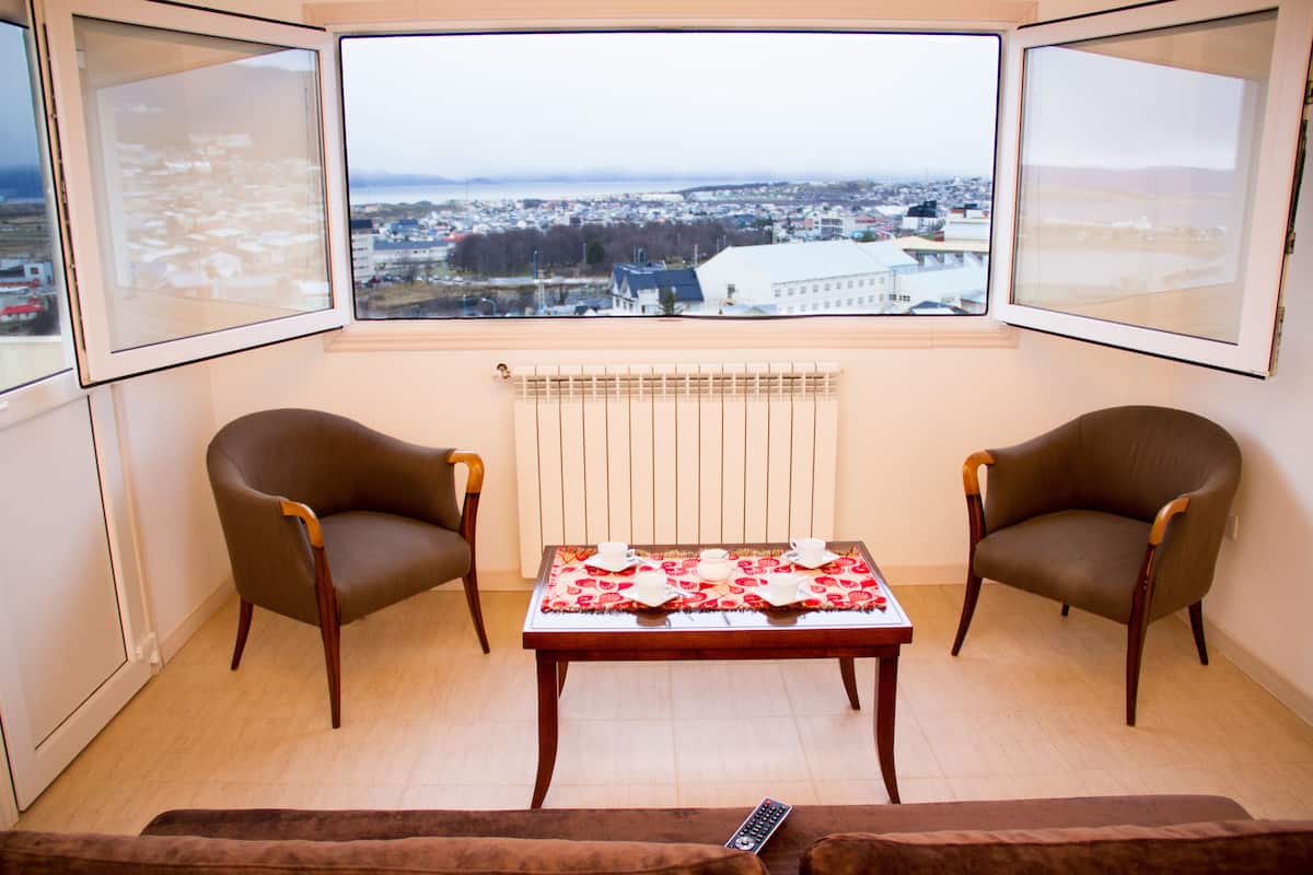 A room with two armchairs, a table set for two, and a view of a cityscape through large windows.