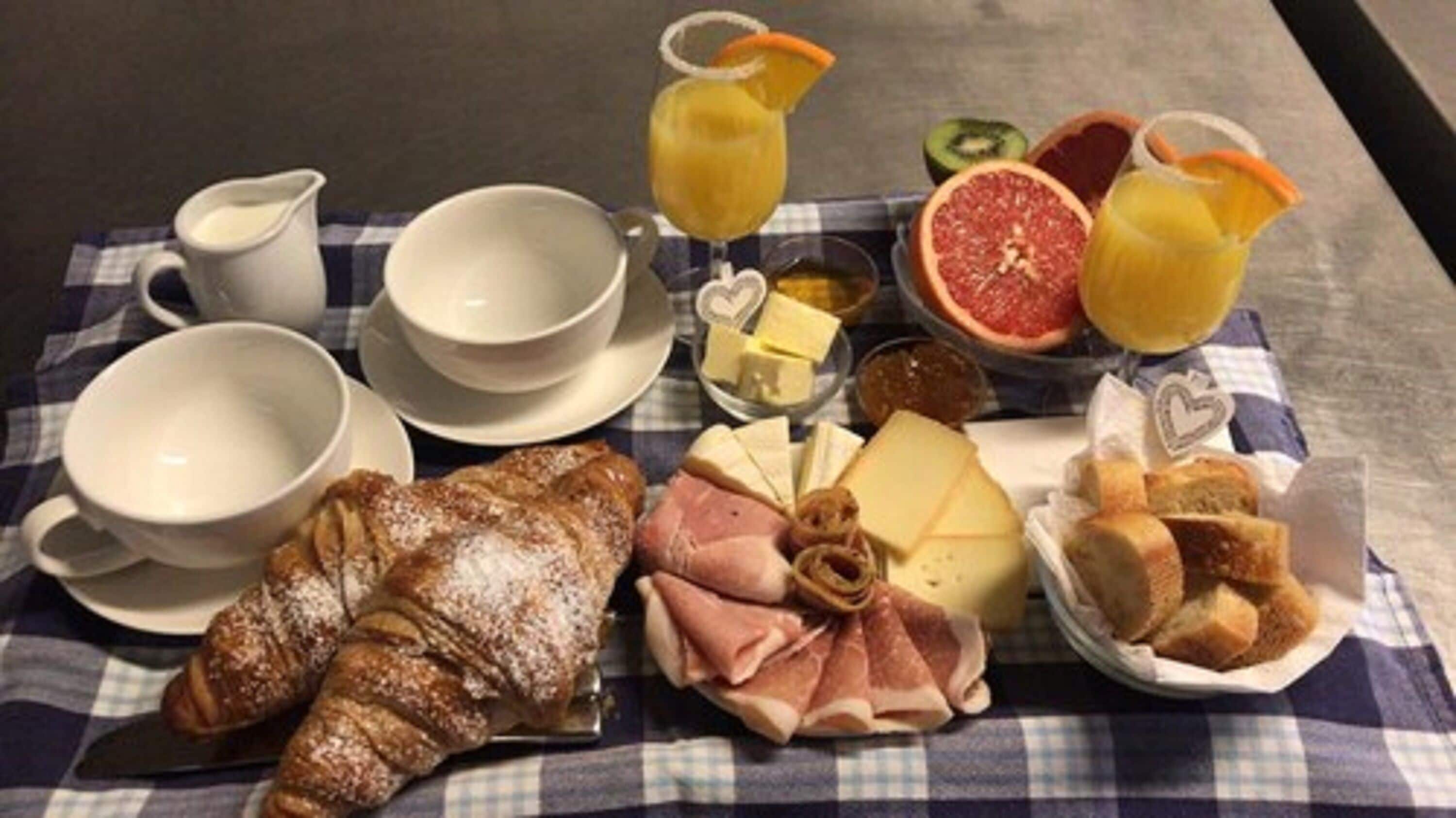 A breakfast setting with croissants, ham, cheese, bread, fruit, and beverages on a checkered tablecloth.